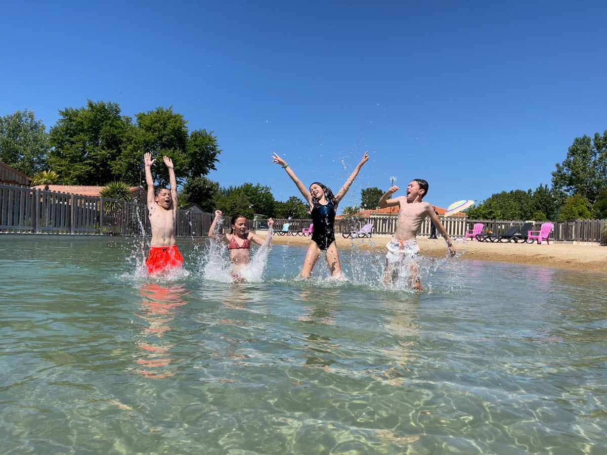 Quatre enfants jouant et éclaboussant dans l'eau claire d'un lagon artificiel avec plage de sable en arrière-plan.
