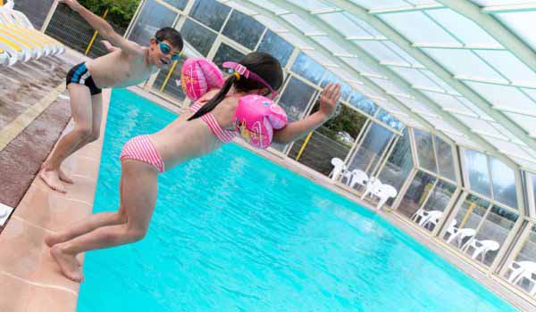 Deux enfants s'amusant à sauter dans l'eau d'une piscine couverte, l'un portant des brassards roses.
